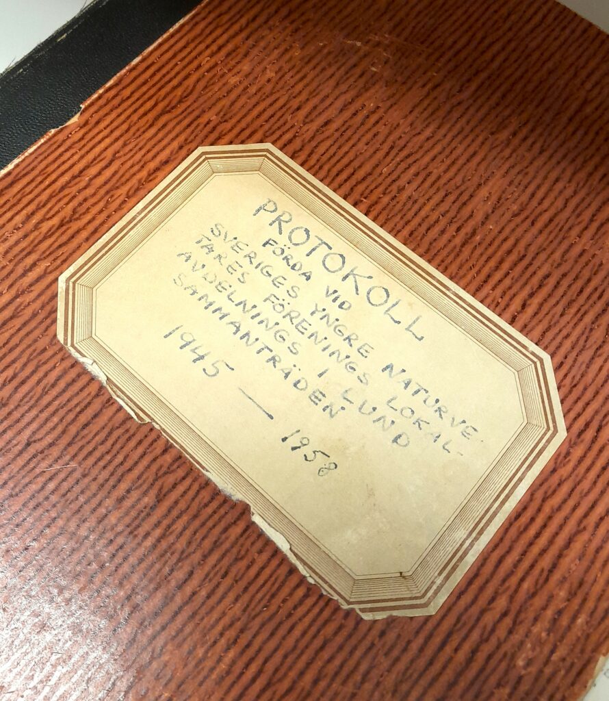 Fotografi av en bok med orange omslag. På en etikett står: "Protokoll förda vid Sveriges yngre naturvetares förenings lokalavdelning i Lund, sammanträden 1945--1958"